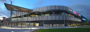 A wide-angle view of Kings Avenue Mall, showcasing its modern architecture, large glass entrance, and busy shoppers.