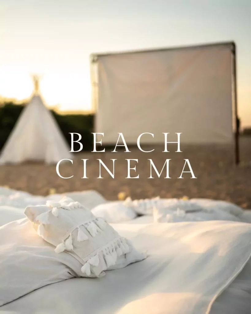 An outdoor beach cinema setup at Sense by the Beach, with a large screen displaying a movie, comfortable seating, and guests enjoying the ocean breeze under starry skies.