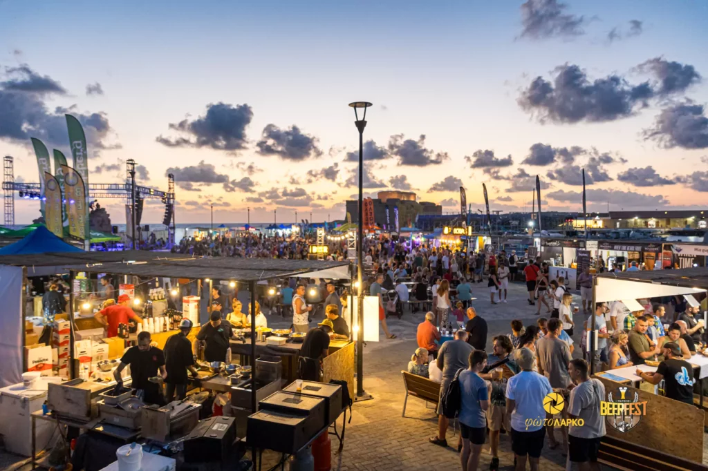 Scenic view of Paphos harbor with the vibrant Paphos Beer Festival setup in the foreground, featuring tents and festivalgoers enjoying the event.