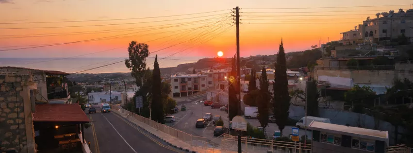 Sunset over a coastal hillside town as a curving road leads downhill past buildings, parked cars, trees, and power lines toward the sea.